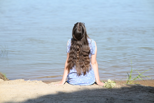 A young girl with long dark hair stands on the bank of the river.