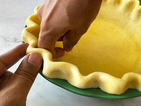 Stock photo showing close-up view of an unrecognisable chef lining a tart tin case with pastry dough by pinching the edge of pie crust with index finger, thumb and knuckle in preparation for baking a pie. Homemade pastry made with white flour and butter ready to be filled with savoury or sweet sauce, ready to bake. Home baking concept.