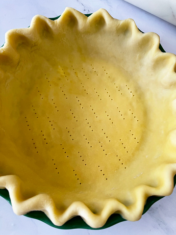 Stock photo showing close-up, elevated view of a freeform fluted, pastry dough lining of a tart tin case that has been pricked with a metal fork, in preparation of baking a pie. Homemade pastry made with white flour and butter ready to be blind baked and filled with a savoury or sweet sauce. Home baking concept.