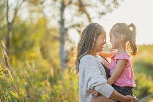 A young Mother of Asian decent holds her daughter up in her arms as they take a walk at sunset on a warm fall evening.  They are both dressed casually and are smiling at one another.