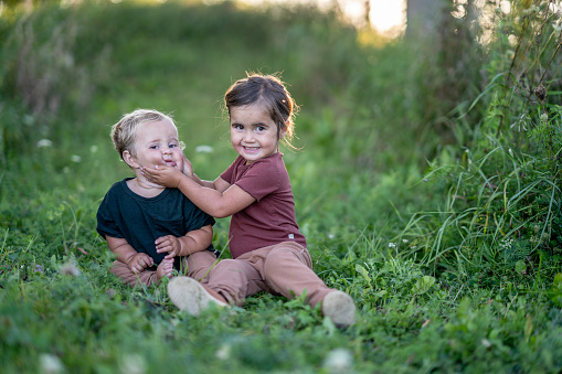 Two sweet young children sit in the grass together as they spend time outside.  The sibling duo is dressed casually and the little girl is squeezing her brothers cheeks gently as she cheekily smiles.