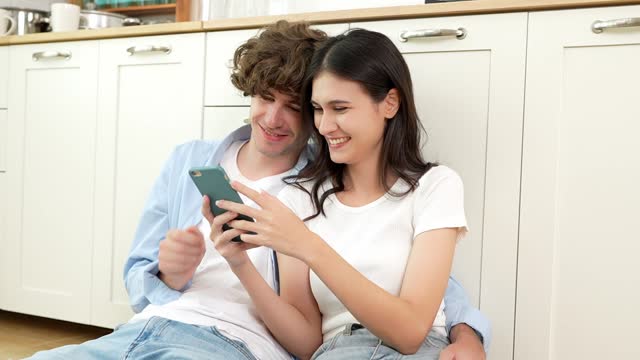 Happy young woman and man using smartphone together boyfriend hug girlfriend sit on floor at home Smiling overjoy wife and husband couple looking at phone screen for shopping online social media