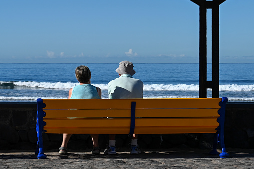 Las Américas, Tenerife, Spain February 12, 2023 -Senior couple sitting on a wooden bench on the beach of Tenerife and looking at the Atlantic Ocean