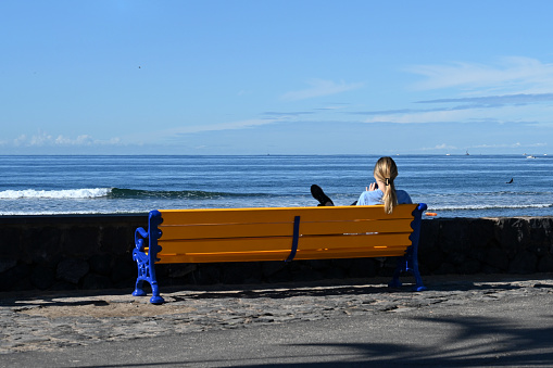 Las Américas, Tenerife, Spain February 12, 2023 -Young lady sitting on a wooden bench on the beach of Tenerife looking into her cell phone.