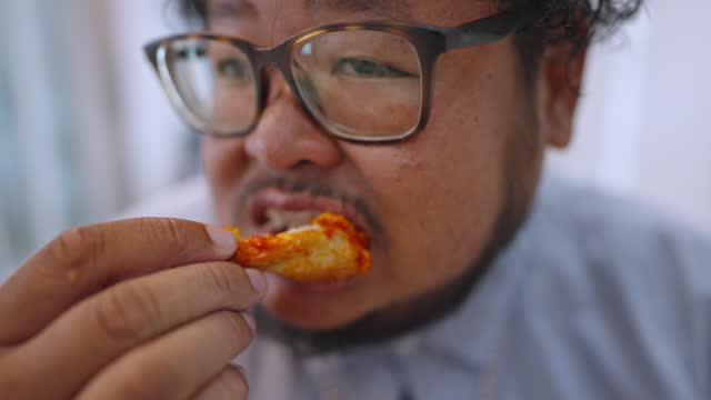 Close-up of a hungry man using his hand eating fried chicken with laughter in his dining room.