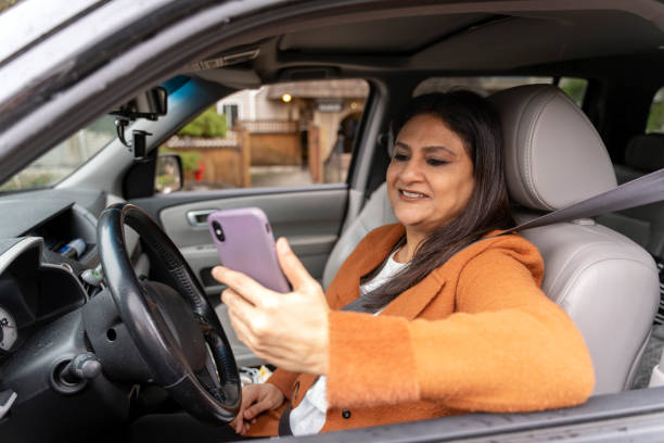 mulher indiana madura sorridente segurando o telefone celular, verificando o e-mail sentado dentro do carro novo - mobile phone seat belt text messaging smiling - fotografias e filmes do acervo