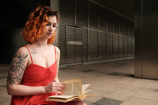 Young caucasian woman reading a book outside. The background is a metallic cosmopolitan one. She is sitting on a bench and wears a red summer dress. Relaxing and peaceful hobbies concept.