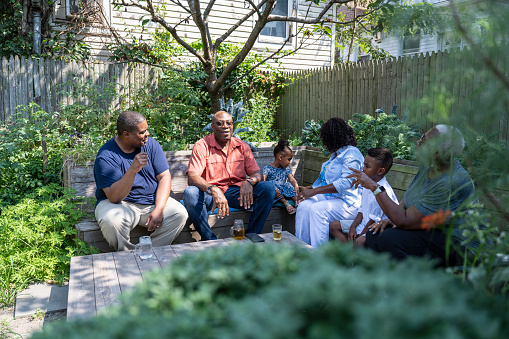 Three generations dressed for summer, sitting together outdoors in Rockaway Beach backyard, surrounded by plants, talking and smiling.