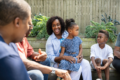 Focus on mature Black woman sitting with young son and daughter, laughing and interacting with multi-generation group in backyard.