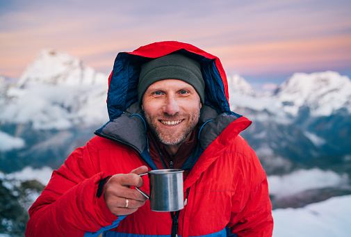 Portrait of smiling at camera high altitude mountaineer dressed red warm dawn jacket holding metal mug of hot tea in with mountains panorama background. Mera Peak High Camp 5700m, Himalayas, Nepal.