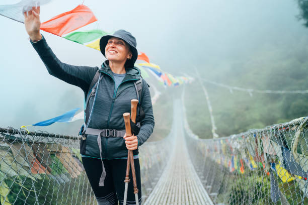 jeune femme souriante traversant le canyon au-dessus du pont suspendu et touchant des drapeaux de prière tibétains multicolores articulés au-dessus de la gorge. trek de la voie d’escalade du pic mera près de lukla, parc national de sagarmatha, népal - lukla photos et images de collection