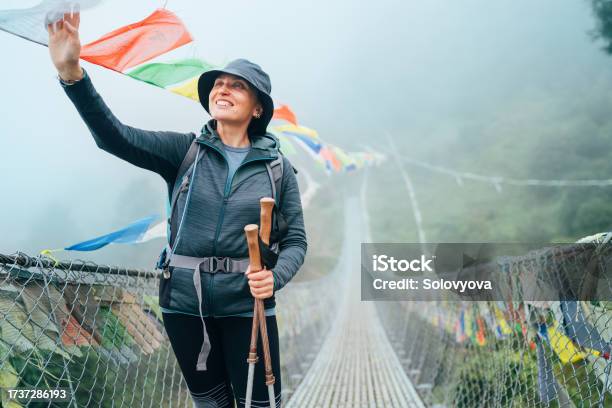 Young Smiling Female Crossing Canyon Over Suspension Bridge And Touching Multicolored Tibetan Prayer Flags Hinged Over Gorge Mera Peak Climbing Route Trek Near Lukla Sagarmatha National Park Nepal Stock Photo - Download Image Now