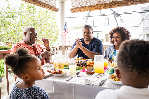 Young Black siblings sitting at dining table on sunny veranda with smiling parents and grandfather, hungry and ready for lunch.