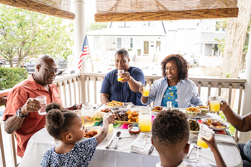 Multi-generation Black family sitting at outdoor table, lifting glasses of lemonade and smiling before beginning summertime feast on veranda.