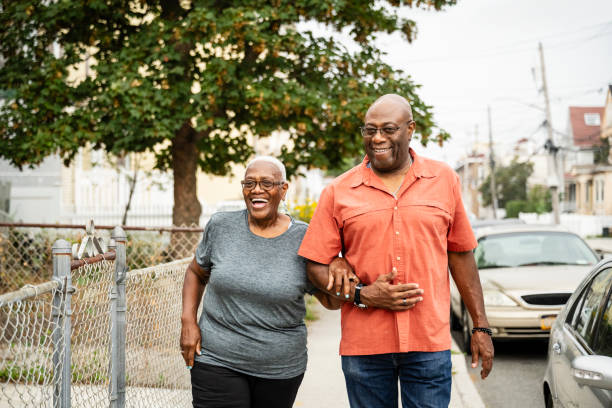 Retired Black couple enjoying exercise in the neighborhood Candid portrait of man and woman, casually dressed and arm in arm, smiling and laughing as they approach camera on sidewalk in Rockaway Beach, Queens. locking-arms stock pictures, royalty-free photos & images