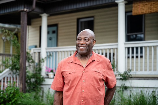 Waist-up front view of balding man in late 60s wearing short sleeved orange shirt, looking away with contented smile, summertime.