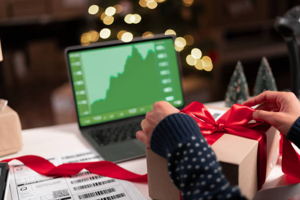 istock Female analyst at her desk works on a laptop showing statistics, graphs while working at Christmas. She Works on the Table packing package gift with red ribbon in creative office. 1737470060