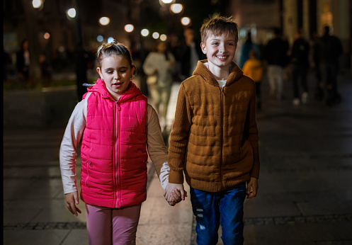 In the captivating ambiance of a city street at night, a brother and sister walk hand in hand, their silhouettes illuminated by the vibrant city lights