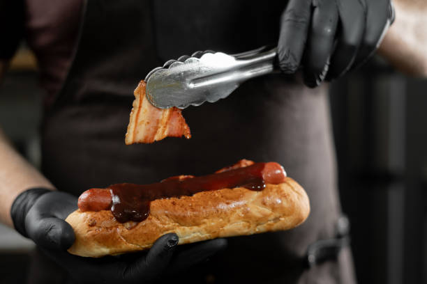 Chef preparing a tasty hot dog with a frankfurter on a fresh roll adding sliced bacon onto sausage Chef preparing a tasty hot dog with a frankfurter on a fresh roll adding sliced bacon onto sausage. Copy space restaurant labor union stock pictures, royalty-free photos & images