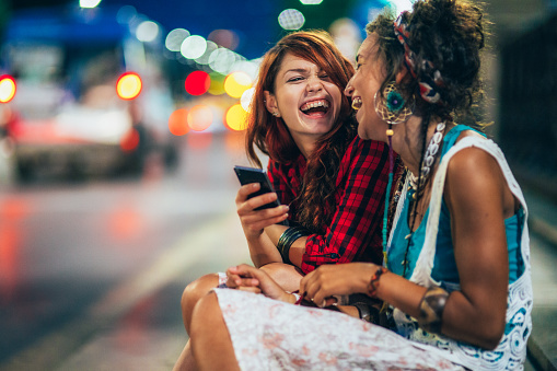 Two young women sitting outdoor in the city and using smartphone. Female friends laughing and talking while texting on the city street at night