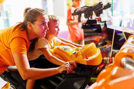 Mother and son play together in an arcade motorcycle simulator in an entertainment center. The concept of family leisure, spending time together
