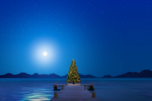 A Christmas tree stands at the end of a pier as a full moon rises over a lake or lagoon on a crystal clear night.