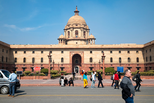 2nd February, 2020 - Delhi, India: This editorial image captures a scene around the government buildings situated in the capital city of Delhi. Known for their unique blend of British colonial and traditional Indian architectural elements, these buildings function as the epicentre for India's legislative and administrative activities. Tourists and local visitors in the photograph are seen taking pictures, exploring the area, and admiring these emblematic structures. Among the buildings featured are the Parliament House and Rashtrapati Bhavan, key sites that not only serve governance functions but also draw attention as important landmarks in the city. The image offers a snapshot of how these governmental edifices intersect with daily life and tourism.