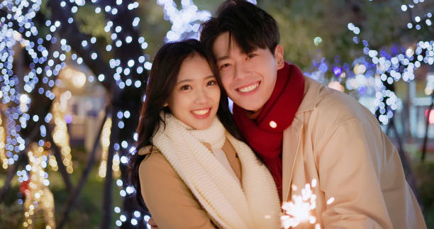 asian happy couple wearing scarves are playing sparklers together at night - looking to you