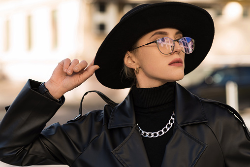 Portrait of an attractive and young blonde woman in a black hat and a leather trench coat while walking around the city. The concept of style and fashion.