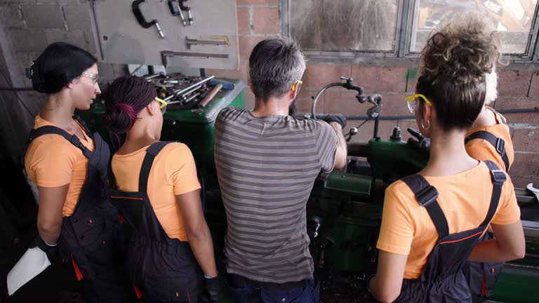 Caucasian male metallurgical engineer, trains group of multiracial female workers how to work on a lathe machine