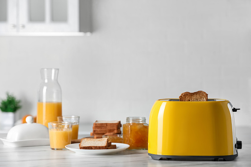 Yellow toaster with roasted bread slices, jam and glasses of juice on white marble table