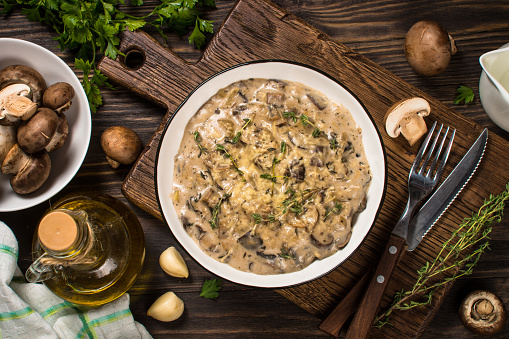 Mushrooms in cream sauce with herbs and cheese. Top view at dark wooden table with ingredients for cooking.