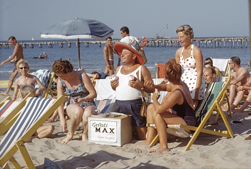 Riccione, Rimini Region, Emilia Romagna, Italy, 1963. Italian ice cream seller in a group of holidaymakers from Central Europe on the beach in Riccione.