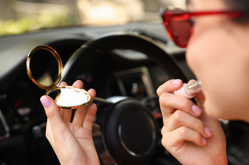 Beautiful woman with cosmetic pocket mirror applying lipstick in car, closeup