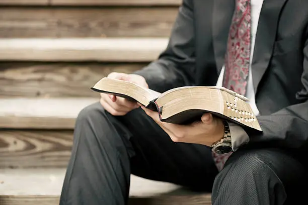A male missionary dressed in a suit sits on wooden stairs and reads from the Scriptures.