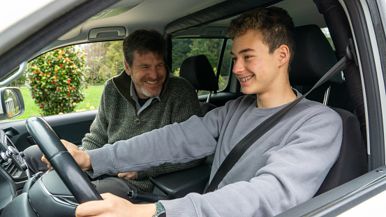 Father teaching his teenage son to drive, both joyfully bonding.