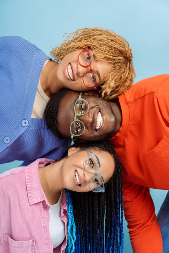 Group of smiling stylish African American friends wearing colorful glasses isolated on blue background. Happy fashion models with trendy hairstyle looking at camera posing for pictures in studio