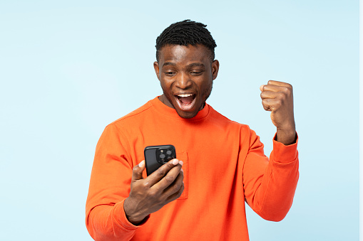 Young overjoyed African American man, gambler holding mobile phone sports betting, win money celebration success isolated on background. Happy guy shopping online with sales. Technology innovation