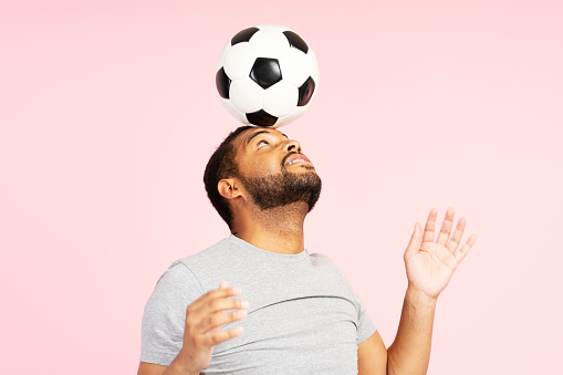 Smiling positive African American young man juggling soccer ball, holding on head isolated on pink background. Attractive young male wearing stylish casual gray t shirt. Hobby concept