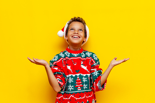cheerful african american boy in christmas clothes holds empty hands and looks up on yellow isolated background, 9 year old child in santa hat dreams and catches copy space