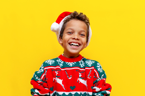 funny african american boy in christmas clothes smiling on yellow isolated background, happy 9 year old child in santa hat laughing