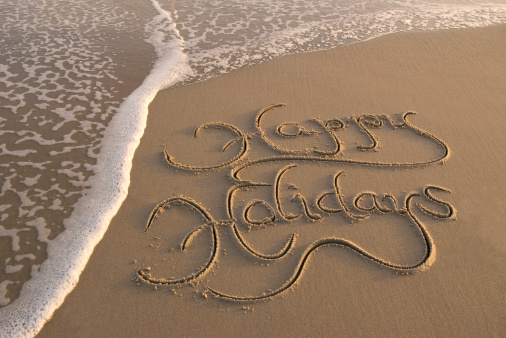Fancy Happy Holidays greeting handwritten message beckons from golden sand next to gentle waves on the beach