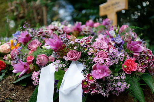 funeral wreath on a grave with white ribbons and flowers in pink tones with a wooden cross in a blurred background