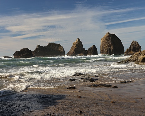 Dramatic scene of waves crashing onto rocky beach with large and awe-inspiring haystack and arch rocks. The sky is a vivid blue with some white clouds. This scene is only visible at low tide which allows access through a tunnel in the surrounding cliffs.  This gives it the name Tunnel Beach in Oregon.