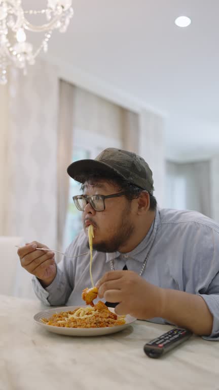 An oversized single man eating Spaghetti Bolognese and fried chicken in a beautiful dining room while watching a soccer match on TV and feeling very happy and satisfied as the team he's cheering won.