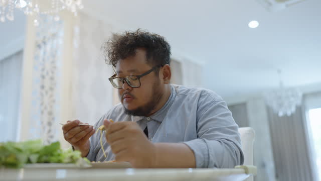 Low angle view of an overweight man eating spaghetti Bolognese with fork and grabbing fried chicken wing with hand and eating it with pleasure in a beautiful dining room.