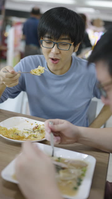 Close-up of two male coworkers talking to each other with happy faces while having crispy mussel omelet and stir-fried noodle in thick gravy as their lunch in a food court at a shopping mall.