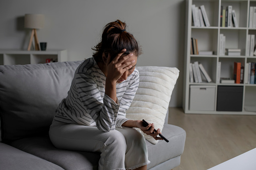Asian woman sitting on sofa holding phone feeling disappointed, sad, upset. Female suffer from mental health problems.
