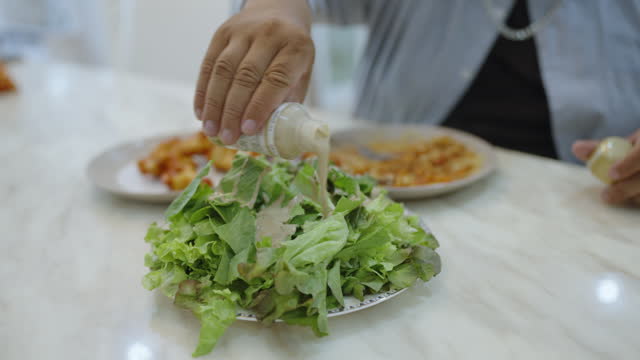 An oversized man pouring cream salad dressing into his green salad bowl and eating green lettuce with fork as an appetizer before having spaghetti Bolognese and fried chicken as main courses while sitting in a dining room.