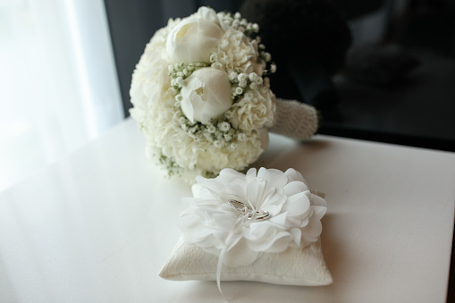 Close-up shot of wedding rings on a silk pillow with white bouquet of fresh flowers on the table